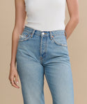A person wearing a white ribbed sleeveless top and high-waisted light blue 90s Jean with hands in pockets, standing against a plain beige background.