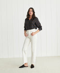 A woman stands indoors against a white paneled wall, wearing a dark plaid shirt tucked into 90's Pinch Waist Jeans in white and black slippers, with one hand in her pocket.