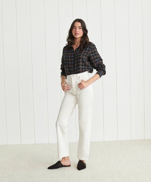 A woman stands indoors against a white paneled wall, wearing a dark plaid shirt tucked into 90's Pinch Waist Jeans in white and black slippers, with one hand in her pocket.