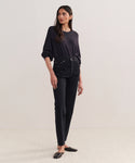 A woman with long dark hair stands against a plain background wearing a loose dark long-sleeve top, black slip-on shoes, and 90s Pinch Waist Long Jeans. She gazes slightly to the side.