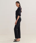 A woman with long, dark hair stands in profile against a light background, wearing a long-sleeve dark top, black flats, and the 90s Pinch Waist Long Jean. Her arms rest naturally at her sides.