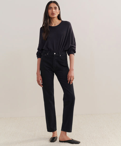 A woman with her hair down and a neutral expression stands against a plain background, wearing a loose black long-sleeve top tucked into 90s Pinch Waist Long Jeans and black slip-on shoes.
