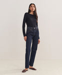 A woman with long, straight hair and a neutral expression stands against a plain background wearing a black long-sleeve top, brown flats, and the 90s Pinch Waist Long Jean in high-waisted dark blue organic cotton.