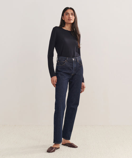 A woman with long, straight hair and a neutral expression stands against a plain background wearing a black long-sleeve top, brown flats, and the 90s Pinch Waist Long Jean in high-waisted dark blue organic cotton.