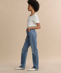 A woman with curly hair stands in profile against a beige background, wearing a white short-sleeve top, 90’s Pinch Waist Long high-rise straight-leg jeans in comfort stretch denim, and white flats, with one hand in her pocket.