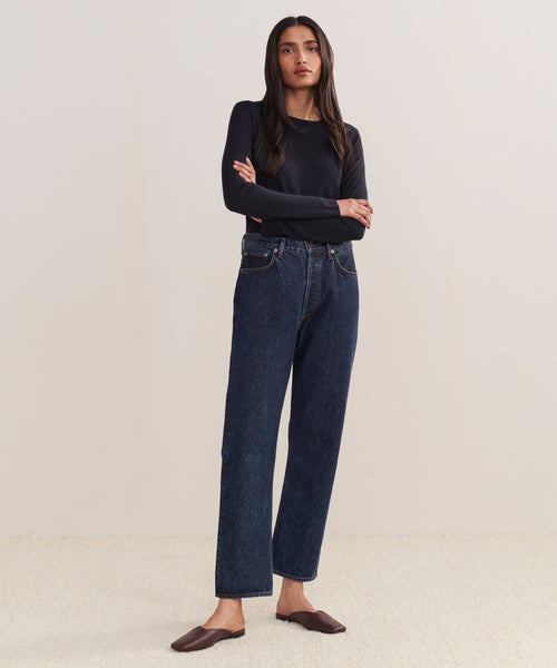 A woman stands with arms crossed, wearing a long-sleeve black top and 90s Pinch Waist Jean against a plain light background.