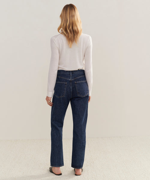 A woman with blonde hair stands facing away, wearing a white long-sleeve top, black shoes, and the 90s Pinch Waist Jean—a high-rise style made from regenerative cotton—set against a plain light background.