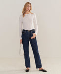 A woman with light hair poses casually against a plain background, wearing a white long-sleeve top, dark blue 90s Pinch Waist Jean, black shoes, and holding a white handbag.