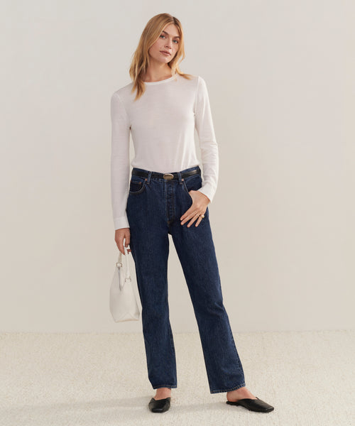 A woman with light hair poses casually against a plain background, wearing a white long-sleeve top, dark blue 90s Pinch Waist Jean, black shoes, and holding a white handbag.
