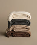 Four Alpaca Basketweave Throws in cream, gray, beige, and brown are stacked against a neutral backdrop. Handwoven in Peru, these throws feature fringed edges and a soft, textured look.