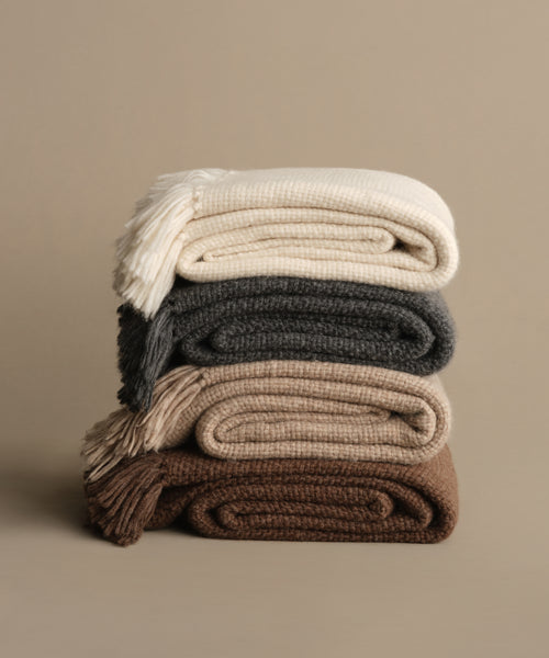 Four Alpaca Basketweave Throws in cream, gray, beige, and brown are stacked against a neutral backdrop. Handwoven in Peru, these throws feature fringed edges and a soft, textured look.