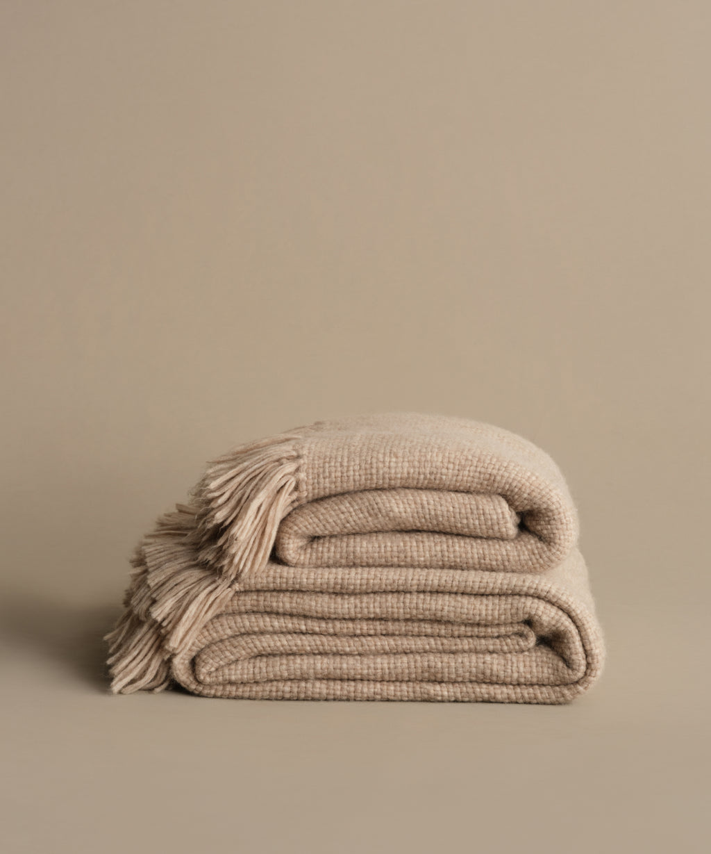 The Alpaca Basketweave Throw, handwoven in Peru from superfine beige alpaca wool with fringed edges, is folded on a matching surface against a plain beige background.