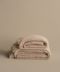 The Alpaca Basketweave Throw, handwoven in Peru from superfine beige alpaca wool with fringed edges, is folded on a matching surface against a plain beige background.