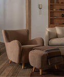 A cozy upholstered armchair in a warm taupe bouclé fabric sits beside a matching ottoman with a striped throw blanket, set in a styled living room with wood flooring and shelving.