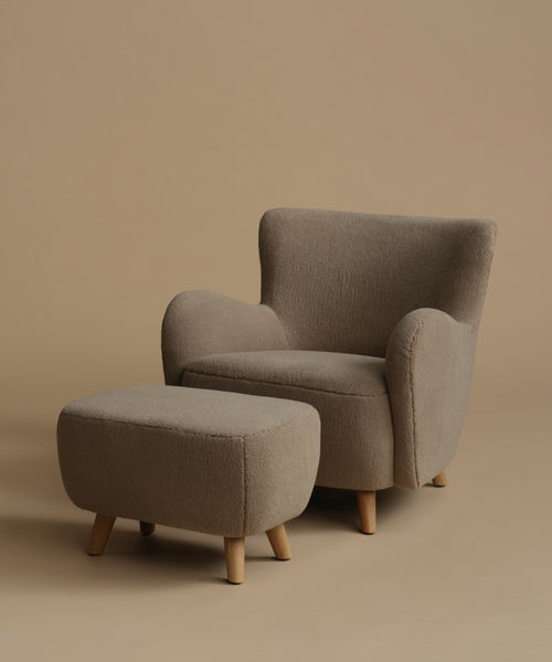 A modern upholstered armchair with a matching ottoman, both in a textured taupe fabric with light natural wood legs, set against a solid warm beige background.