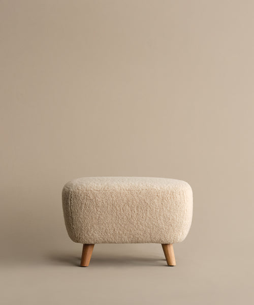 The Aspen Ottoman, a small, light beige piece with textured Sherpa fabric and wooden legs, sits against a plain beige background.