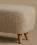 A low, square upholstered ottoman stool with a textured bouclé fabric in a light beige color and four tapered wooden legs, set against a solid warm beige background.