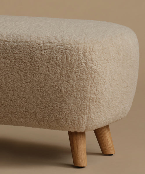 A low, square upholstered ottoman stool with a textured bouclé fabric in a light beige color and four tapered wooden legs, set against a solid warm beige background.