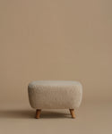 A low, square upholstered ottoman stool with a textured bouclé fabric in a light beige color and four tapered wooden legs, set against a solid warm beige background.