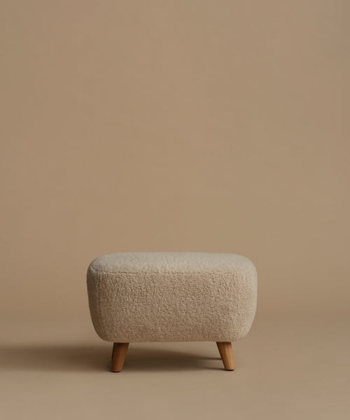 A low, square upholstered ottoman stool with a textured bouclé fabric in a light beige color and four tapered wooden legs, set against a solid warm beige background.