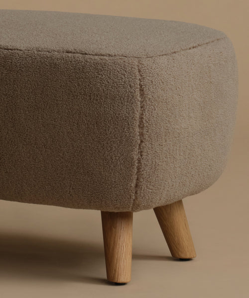 A close-up of a round upholstered ottoman  with a textured bouclé fabric in a warm taupe-beige color, supported by four tapered light wood legs, set against a solid beige background.