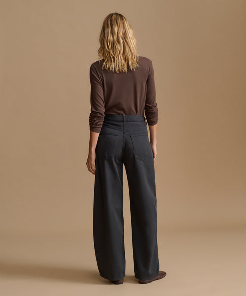A woman is shown from behind wearing a dark brown long-sleeve top and dark charcoal gray wide-leg trousers, standing against a solid beige studio background.