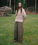 A woman stands on a grassy lawn wearing a cream knit sweater with a brown geometric Fair Isle pattern and wide-leg olive green pants, with a forest and wooden structures in the background.