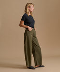 A woman stands in profile wearing olive green wide-leg trousers, a black short-sleeve t-shirt, and black flats against a solid beige studio background.