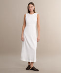 A woman stands against a beige background wearing the sleeveless, ankle-length Blair Dress in white linen—an ideal spring piece. She pairs it with black woven flats, her straight brown hair framing her neutral expression.
