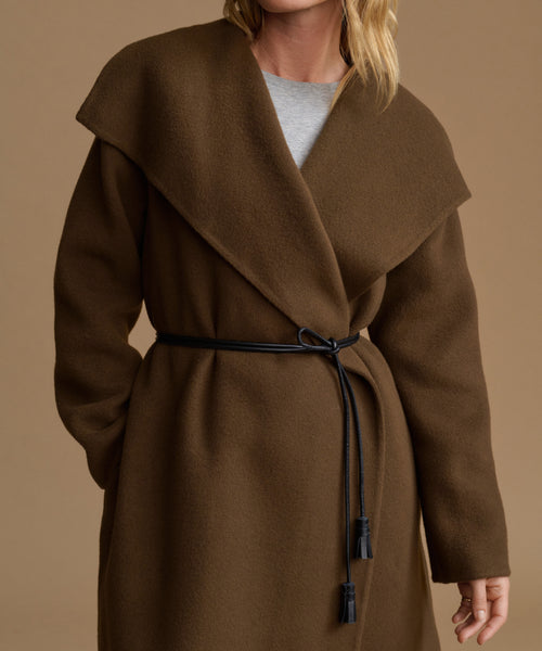 A woman wearing a brown wrap coat with a large shawl collar and a black belt, shown against a plain beige studio background.