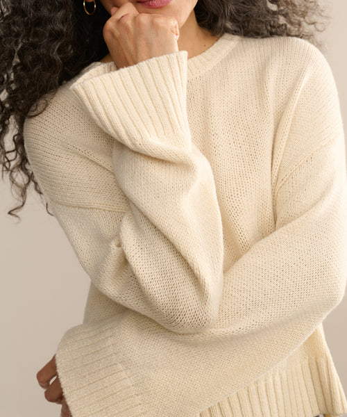 A woman with curly hair wears the cream-colored, oversized Cameron Crewneck. Her hand near her face draws attention to the textured fabric and roomy sleeves, set against a neutral background.