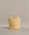 The Capri Bag is a handcrafted woven straw tote with two short white handles, shown against a plain neutral background.