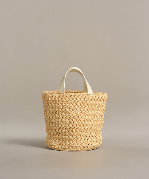 The Capri Bag is a handcrafted woven straw tote with two short white handles, shown against a plain neutral background.