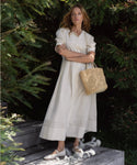 A woman stands outdoors among green bushes, wearing a long cream dress, white flats, and holding the Capri Bag. She has long light brown hair and looks at the camera with folded arms, radiating Tuscany-inspired charm.
