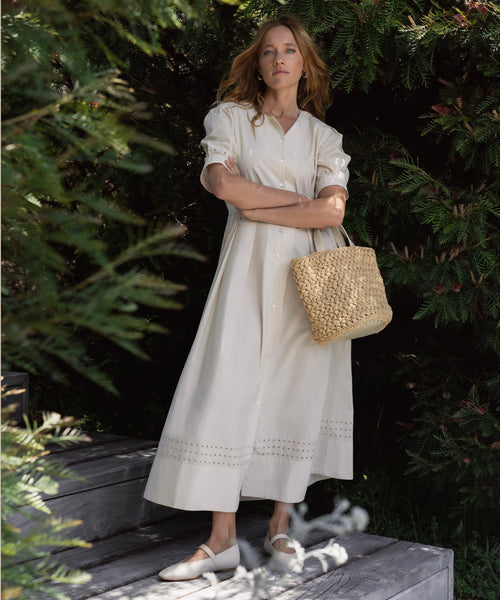 A woman stands outdoors among green bushes, wearing a long cream dress, white flats, and holding the Capri Bag. She has long light brown hair and looks at the camera with folded arms, radiating Tuscany-inspired charm.