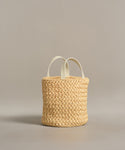 The Capri Bag is a handcrafted woven straw basket with two short white handles, displayed on a plain light gray background.