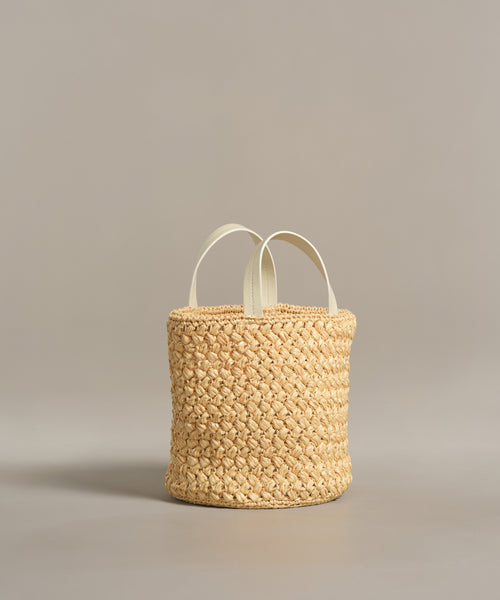 The Capri Bag is a handcrafted woven straw basket with two short white handles, displayed on a plain light gray background.
