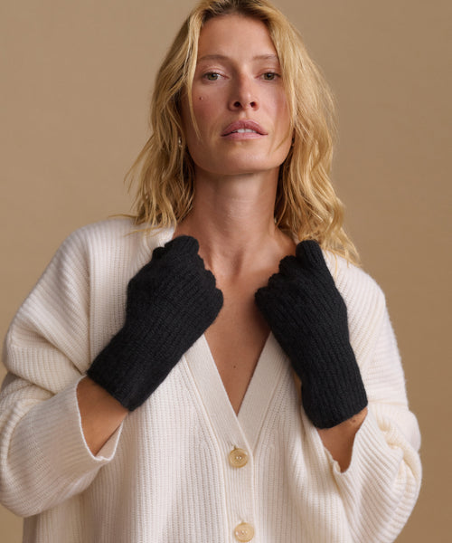 A woman wearing black knit fingerless gloves over a cream ribbed cardigan, posed against a solid beige background.