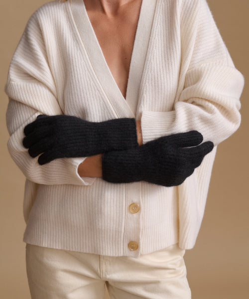 A person wearing a cream ribbed cardigan and cream pants, with black knit gloves crossed over the chest, against a warm beige studio background.