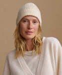 A woman wearing a cream ribbed knit beanie and a matching cream knit top, photographed against a warm beige studio background.