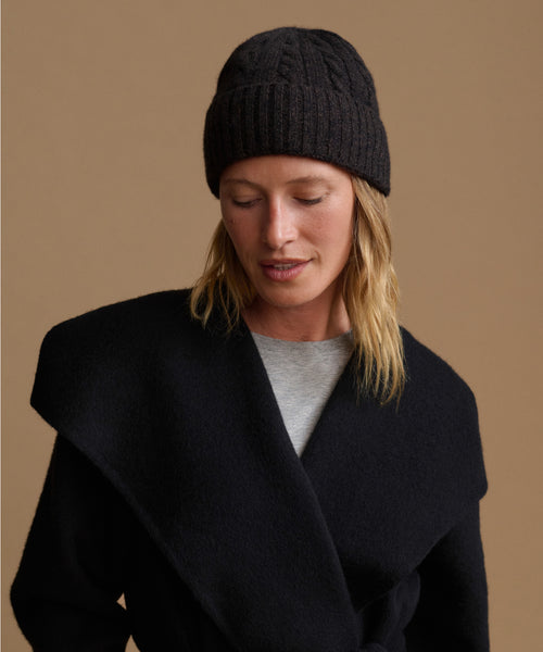 A woman wearing a black cable-knit beanie and a black wrap coat over a light gray top, photographed against a warm tan studio background.