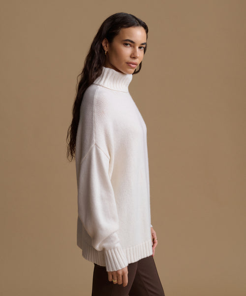 A woman wearing a cream-colored ribbed turtleneck sweater, shown in profile against a solid tan studio background.