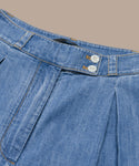 Close-up of the waistband and upper section of the Denim Newport Trouser in blue Italian denim, featuring two white buttons, orange stitching, and pleated detailing on a beige background—ideal for spring and summer.