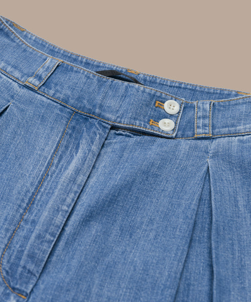 Close-up of the waistband and upper section of the Denim Newport Trouser in blue Italian denim, featuring two white buttons, orange stitching, and pleated detailing on a beige background—ideal for spring and summer.