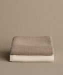 A Chateau Alpaca Coverlet in ivory is neatly folded and stacked atop a beige blanket against a plain, light brown background.