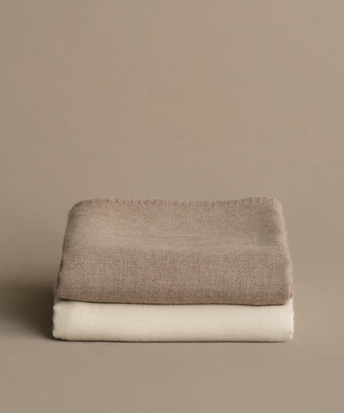 A Chateau Alpaca Coverlet in ivory is neatly folded and stacked atop a beige blanket against a plain, light brown background.