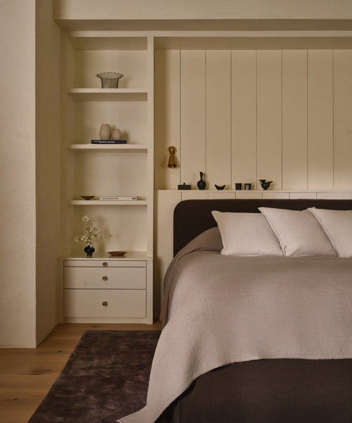 A cozy bedroom features a neatly made bed with white pillows and a Chateau Alpaca Coverlet. Built-in shelves beside the bed showcase books, vases, and decor. The room embraces warm, neutral tones and minimalist style.