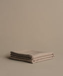 The Chateau Alpaca Coverlet, folded in beige, rests on a matching surface against a plain beige background, creating a minimalistic and neutral scene.