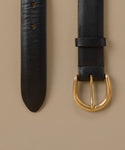 A close-up of a black leather belt laid flat on a warm beige background, showing the textured strap, adjustment holes, and a polished gold-tone metal buckle.