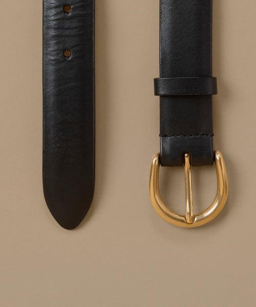 A close-up of a black leather belt laid flat on a warm beige background, showing the textured strap, adjustment holes, and a polished gold-tone metal buckle.